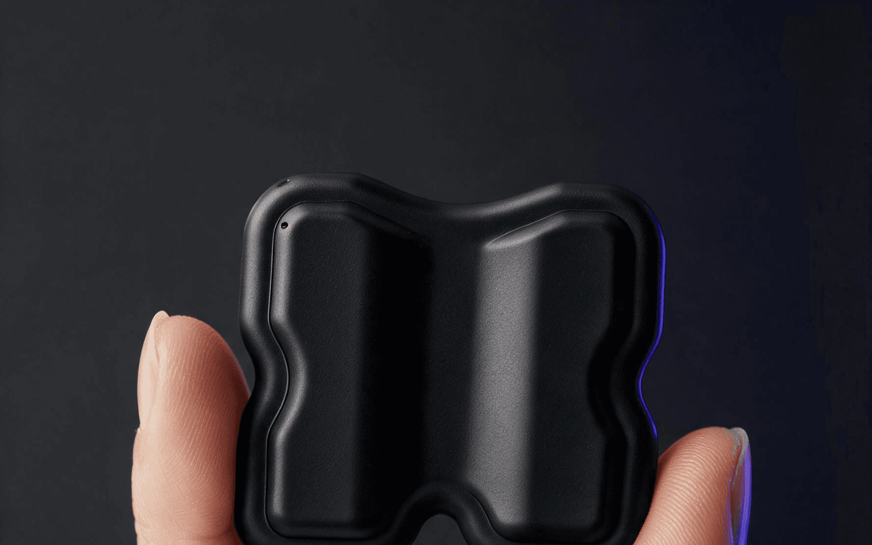 Fingers holding the KTRL patch: H-shaped black conductive silicone single-use patch pinched between thumb and forefinger